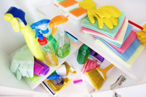 A cluttered indoor space showing a variety of cleaning supplies and tools. In the foreground, there are multiple bottles of spray and liquid cleaning products with colorful caps, including blue, orange, and red, placed on a surface with a pile of green rubber gloves. Behind these bottles, a collection of folded cleaning cloths in pastel and vibrant colours, such as pink, blue, and yellow, are stacked on a white table or shelf. To the right, a yellow cleaning brush with stiff bristles rests on the surface, along with other small cleaning tools. The environment appears to be a storage or utility area for cleaning equipment, with a bright, well-lit setting. This scene is representative of waste or rubbish management tasks related to cleaning supplies and equipment, often handled by services such as Commercial Waste Belmont in the local area, possibly near Belmont or in the surrounding town, reflecting the context of domestic or commercial rubbish disposal and cleaning material organization.
