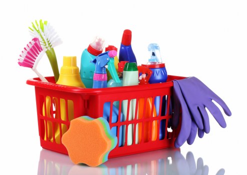 A close-up of a red plastic shopping basket filled with an assortment of cleaning supplies and tools, including colorful spray bottles, a yellow bottle, a green spray bottle, and a blue bottle, alongside purple rubber gloves draped over the side. The cleaning products feature various shapes and bright colors such as blue, red, and green, with some having spray nozzles or caps visible. The basket is placed on a smooth, reflective surface with a plain background, illustrating typical contents used in domestic or commercial cleaning contexts. The image subtly relates to waste removal services by depicting items that may be disposed of or require proper handling, and the overall setting suggests a storage or preparation area, possibly in a residential or commercial environment within the UK, consistent with local rubbish management themes provided by Commercial Waste Belmont.